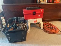 Basket of assorted hand tools including jumper cables, files, with a red plastic tool box on a white plastic stool and an orange extension cord coiled beside.
