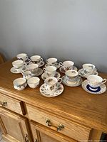 Full view of 10 assorted vintage cups and saucers displayed on wooden sideboard with 3 serving pieces including creamer, sugar bowl, and small jug.
