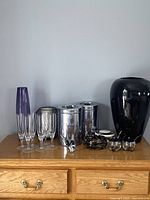 View of all 15 items on a wooden dresser: 6 crystal glasses, tall purple glass vase, black metal candle holder, black art glass bowl, glass condiment dishes with lids, large black ceramic vase, retro lidded ceramic dish, art glass bird, and 2 stainless steel Garnerware canisters with visible wear.