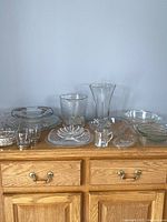 Full view of the 17 pieces of clear glassware laid out on a wooden dresser showing all items including plates, bowls, vase, ice bucket, and dishes.