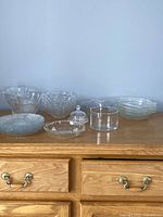 22 pieces of clear vintage glassware including fish plates, flower plates, small bowls, divided dish, candle holders, and butter bell displayed on wood surface.