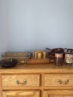 Full lot overview showing all 8 items arranged on a wooden dresser: clear glass baking dish in a wicker tray, two brown glass oven dishes, vintage wooden rolling pin, retro wooden salt/pepper/napkin holder, wooden salad bowl set with tongs, and two ceramic pitchers.