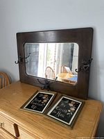 Full view of the antique solid wood framed mirror with visible cast iron hooks on all sides, placed on a wooden cabinet. Below it are two framed foil prints.