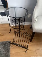 Photo showing the black metal round bistro table with glass top and metal floral pattern lower shelf next to a beige upholstered chair, and the black metal magazine rack placed on the floor in front.