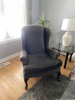 Brown fabric wingback-style chair with curved armrests and dark wooden legs on hardwood floor next to a glass side table with lamp and a plant in the background.