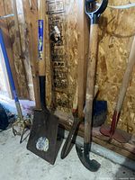 Photo showing a flat-edged shovel, a hoe and other gardening tools leaning against a wooden wall inside a shed