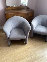 Two grey fabric retro tub chairs in front of a wooden dresser, showing full view of chairs and upholstery.
