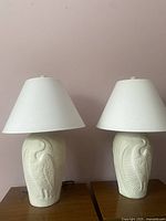 Pair of tall white ceramic lamps with embossed crane motifs and white shades displayed side by side on wooden surfaces against a pale pink wall.