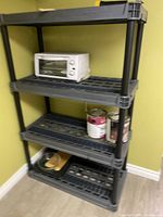 Tall black plastic shelving unit with four open grid shelves, used for storage, contains items but contents not included.