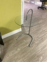 Small round glass side table with clear glass top and curved metal frame, placed on wood floor against yellow wall.