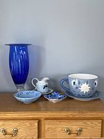 Set of five decorative items displayed together on a wooden surface: tall blue ribbed glass vase, small blue bowl, Ames water pitcher and matching bowl, small pansy plate, and a large blue teacup planter with saucer.