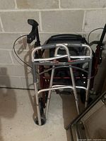 Two walking devices positioned close together in a basement or storage area, showing the silver two-button walker in front of the maroon rollator.