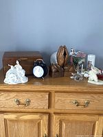 Vintage home decor items on wooden dresser: wooden box, twin bell alarm clock, ceramic figurines, bookends, glass candle holder, vase, decorative plate.