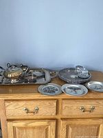 Full view of vintage silver plated tea service set on large rectangular tray along with round tray containing two glass condiment dishes and three decorative plates arranged in front.