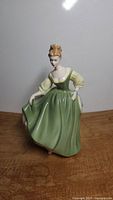 Full front view of the Royal Doulton Fair Lady porcelain figurine wearing a long green dress and cream puffed sleeves on wooden surface.