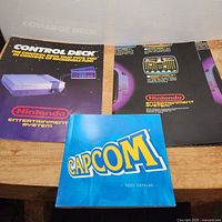 Photo showing the 2003 Capcom catalog in front of the Nintendo manual and poster spread on a wooden surface.