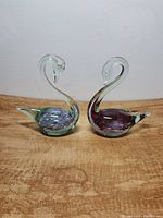 Pair of vintage glass swans side by side on a wooden surface, showing pink and purple coloration with clear glass heads and necks.
