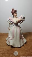 Full front view of the Lenox hand painted porcelain lady figurine in Victorian gown holding an open book or fan, next to a coin for scale.