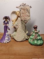 Photo showing three figurines: Enesco angel with metal sign, green dress Josef Originals figurine, purple dress Thomas Kinkade Victorian figurine.