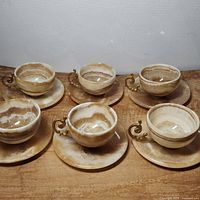 Photo showing six onyx teacups with gold color ornate handles arranged with matching saucers on a wooden surface.