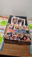 Closed box of NBA Hoops basketball cards on a table alongside NBA Bip Quiz books. Box features colorful fan and player graphics from the 1990s.
