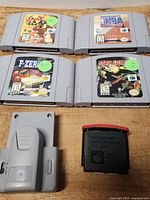 Four Nintendo 64 game cartridges and two gaming accessories laid out on a wooden surface: Blast Corps, Kobe Bryant in NBA Courtside, F-Zero X, Star Soldier: Vanishing Earth; plus a gray Memory Expansion Pack and a gray Rumble Pack.