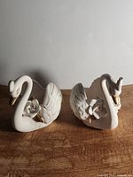 Two white ceramic swan planters with gold accents and rose details, positioned side by side on a wooden surface against a plain wall.