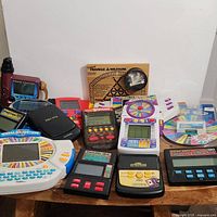 Photo of 14 vintage pocket electronic games laid out on a white surface showing various designs and colors.