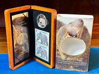 Open wooden case displaying the coin, two stamps, and certificate, alongside the protective sleeve with grizzly bear image.