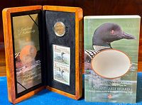 Wooden display case open to show the coin, stamps, and certificate of authenticity with the outer illustrated sleeve beside it.