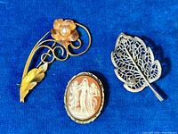 Set of three antique brooches displayed on a blue cloth background showing the gold filled flower brooch with pearl, carved cameo on silver, and filigree leaf brooch.
