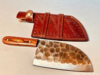 Top-down view of the chef cleaver and its brown leather sheath, showing the textured hammered pattern on the blade and polished wood handle with brass rivets.