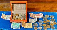 Wide shot of coins in wooden box and various banknotes and coins spread on blue cloth.