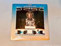 Front cover of 'All The World's A Stage' double vinyl LP showing stage setup with drum set and illuminated star man logo.