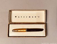 Top view of the Waterman fountain pen inside original box showing the black pen body and gold-tone cap with three rings, and the interior lid with 'Waterman's' printed.
