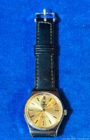 Top view of vintage CAMY Geneve wristwatch showing gold-tone dial and black leather strap on blue background.