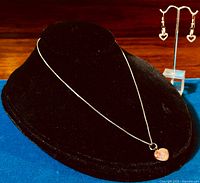 Sterling silver box chain necklace with pinkish art glass heart pendant displayed on a black velvet display stand. Pair of sterling silver heart-shaped earrings shown hanging on a clear acrylic stand.