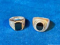 Two vintage men's rings lying on blue fabric: one with rectangular onyx stone and engraved band, the other with oval onyx stone and Greek key bezel.