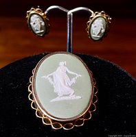 Front view of large oval Wedgwood jasperware pendant/brooch with white relief classical figure on pale green background and gold-filled scalloped edging; matching earrings visible behind on stand.