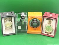 Four wristwatches in original or display packaging, two analog and two digital, arranged in a row against a green background.