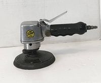 Side angled view of the Sata dual action pneumatic sander showing logo sticker, ergonomic black rubber handle, and metal body with sanding pad.