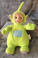 Front view of green Dipsy Teletubby plush talking doll showing soft fabric body, plastic face, blue square on front and antenna on head.