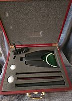 Open wooden case showing a black metal putter, two white golf balls, a green and white ball holder, and three golf tees arranged in foam cutouts