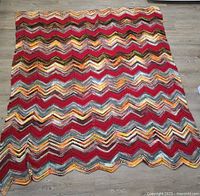 Photo shows a handmade knitted afghan blanket spread out on a floor, featuring a chevron zigzag wave pattern in red, yellow, brown, cream, and orange colors.