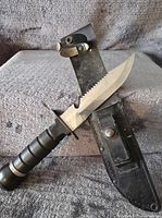 Rambo style hunting knife lying across a black sheath, showing serrated edge and black handle grip.