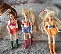 Three vintage Sailor Moon character figures lying on a gray fabric surface. Each figure wears a different colored sailor-style costume with matching boots and gloves, with worn and messy hair tied back.