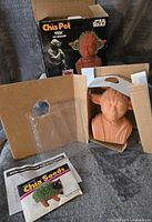 Opened box with Yoda chia pet planter and seed packet inside on a grey surface.