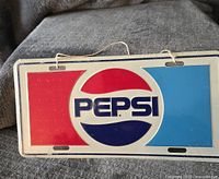 Front view of vintage Pepsi-Cola metal license plate with red, blue, and white Pepsi logo and string attached.