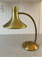 Front and side view of brass coloured table lamp with conical shade, flexible neck, and round base on a light colored counter against white tiled wall.