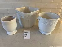 Photo showing all three ceramic and pottery containers on a kitchen counter: a hexagonal container with wing motifs, a round container with fan patterns and handles, and a small pedestal cup.